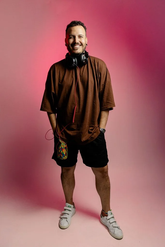 Homem sorrindo, usando camiseta marrom, short preto, tênis branco, com fone de ouvido em volta do pescoço e um estojo colorido pendurado no bolso, em fundo rosa.