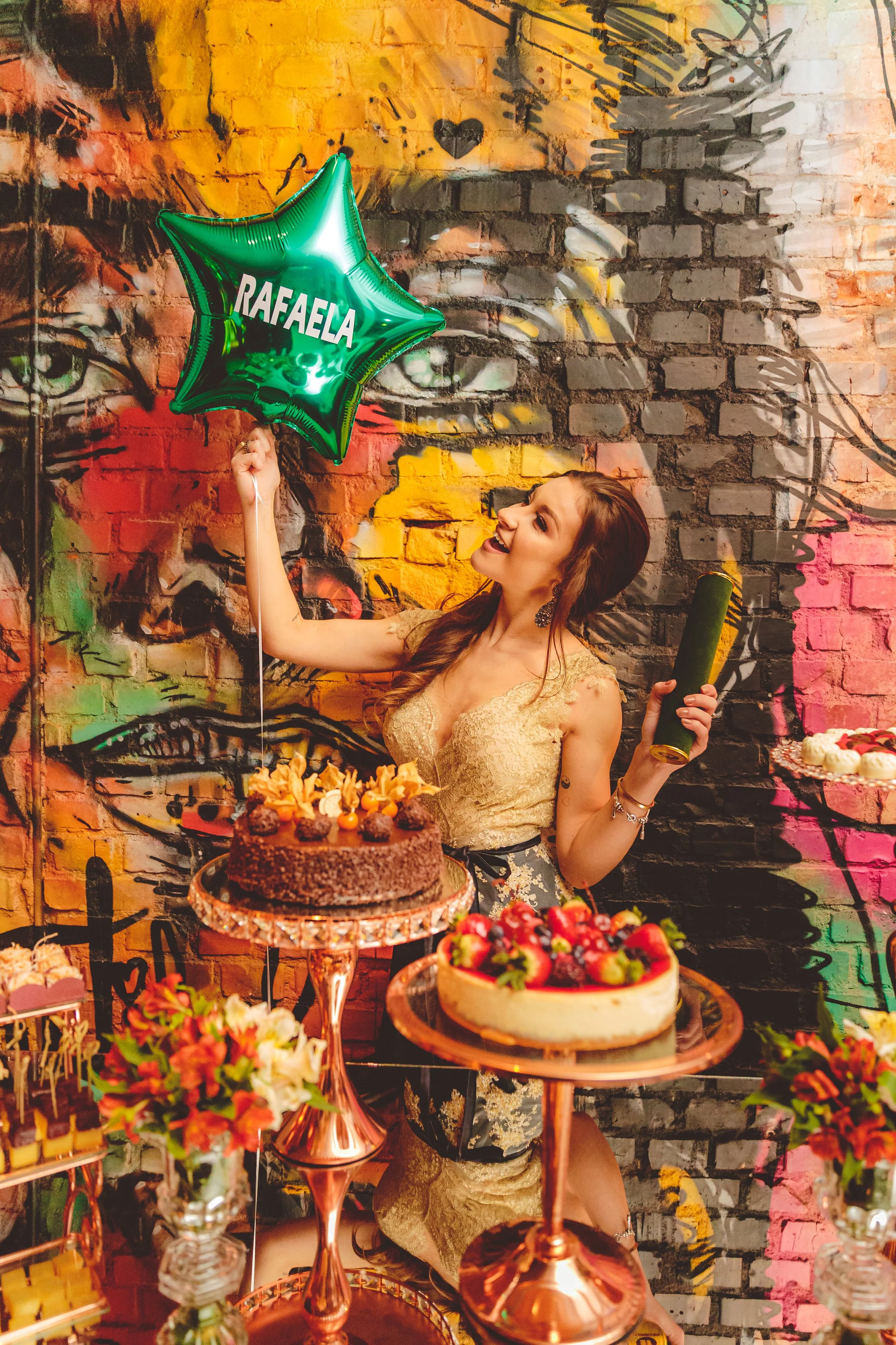 Jovem sorridente segurando um balão verde em formato de estrela com o nome 'Rafaela', ao lado de um bolo de aniversário com frutas e decoração festiva, em um ambiente decorado com grafite em uma parede de tijolos.