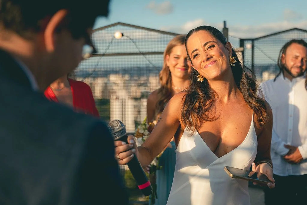 Mulher sorridente segurando um telefone e microfone durante um evento ao ar livre, com várias pessoas ao fundo, sob céu claro.
