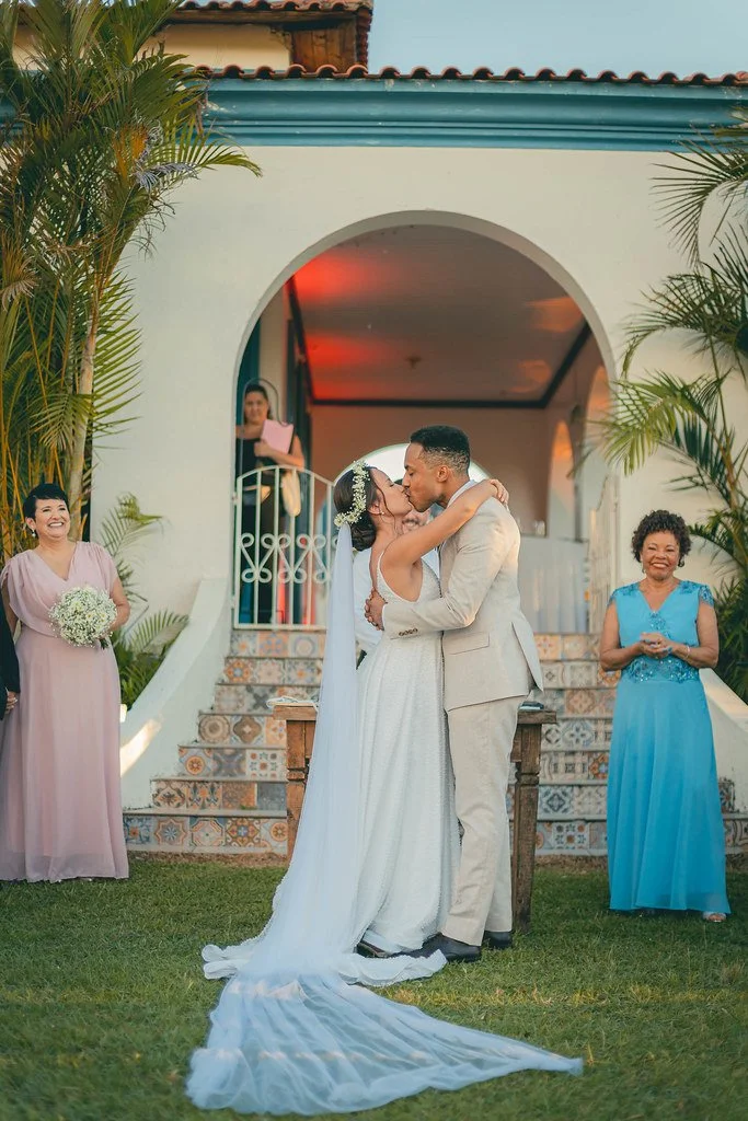 Casal de noivos se beijando no altar ao ar livre, cercado por familiares sorridentes, durante uma cerimônia de casamento.