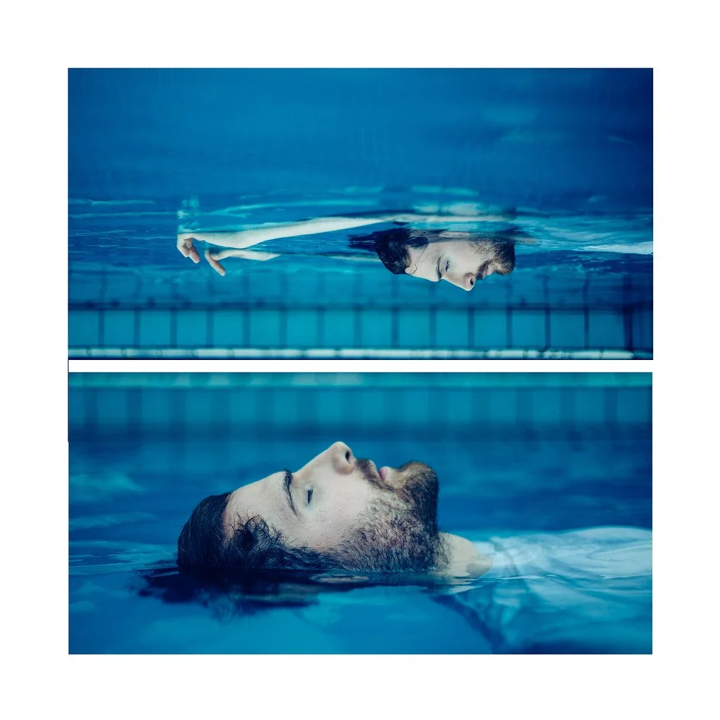 Homem com barba e cabelo escuro, de olhos fechados, está deitado na água de uma piscina, com a cabeça parcialmente submersa. Acima dele, na mesma piscina, há uma mulher com cabelo escuro, de perfil, também de olhos fechados, com um braço estendido na