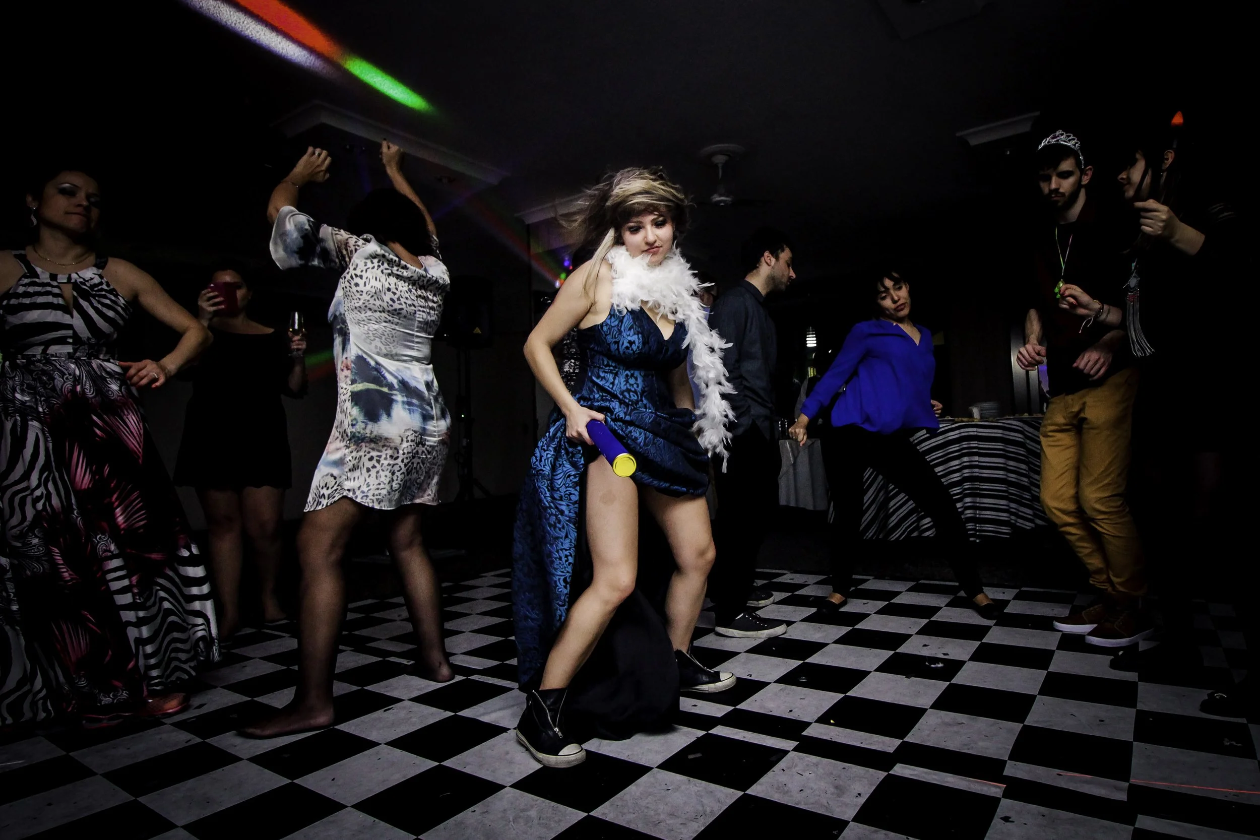 Pessoas dançando em uma festa, com uma mulher no centro usando uma roupas azul, uma faixa de pena branca no pescoço e um microfone na mão, no ambiente com piso quadriculado preto e branco e luzes coloridas.