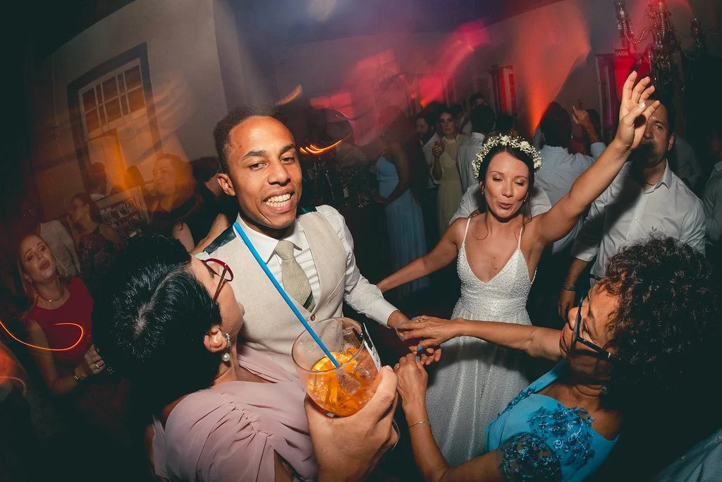 Pessoas dançando em uma festa, incluindo um homem com terno e uma mulher com vestido branco e tiara, em uma celebração animada com iluminação colorida.