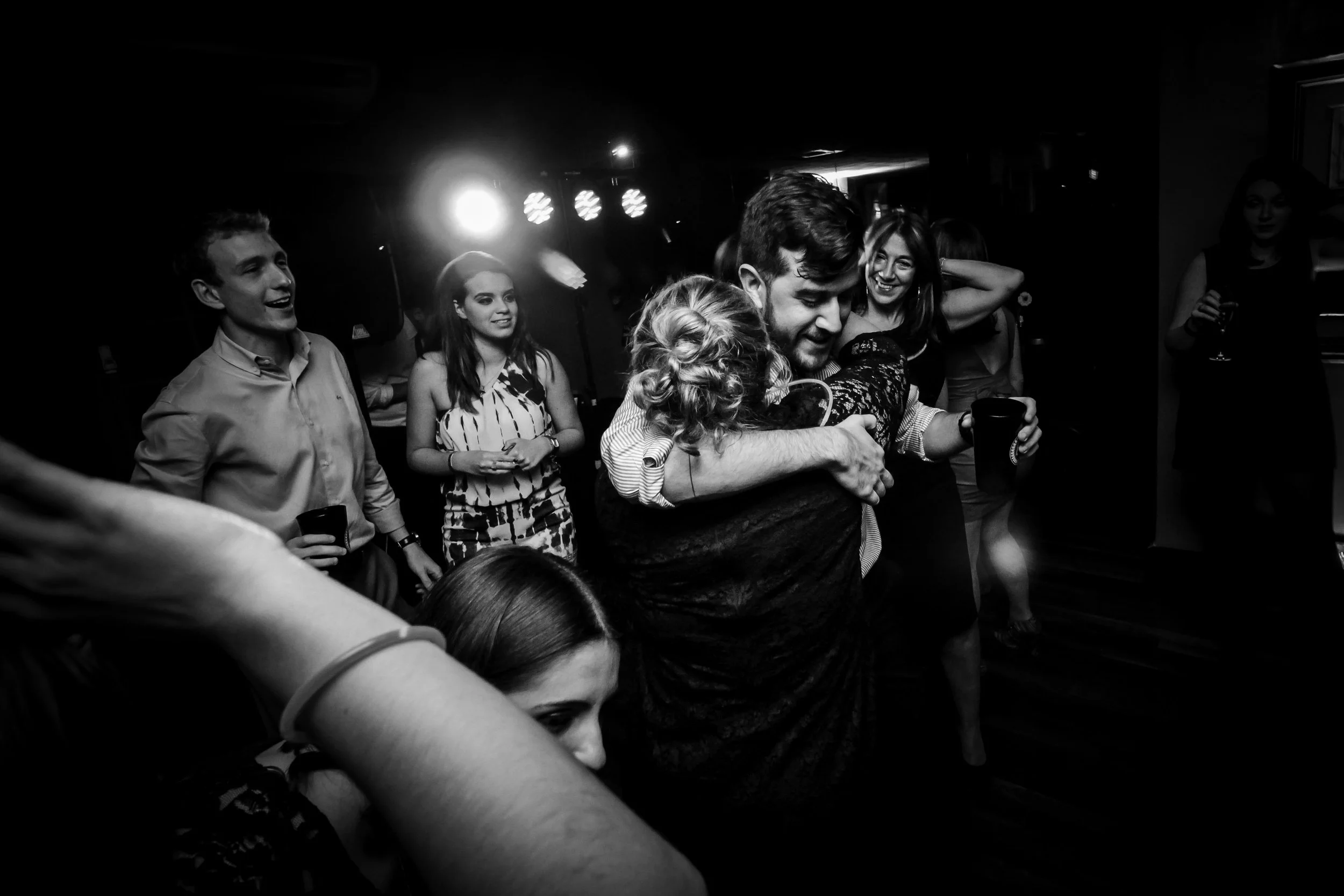 Pessoas celebrando e se abraçando em uma festa, com iluminação de palco ao fundo, em ambiente interno.