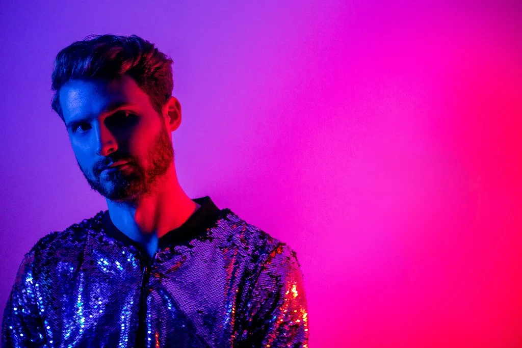 Homem com cabelo e barba escuros, vestindo jaqueta de paetês brilhantes prateados, em iluminação de luz azul e rosa, contra fundo rosa