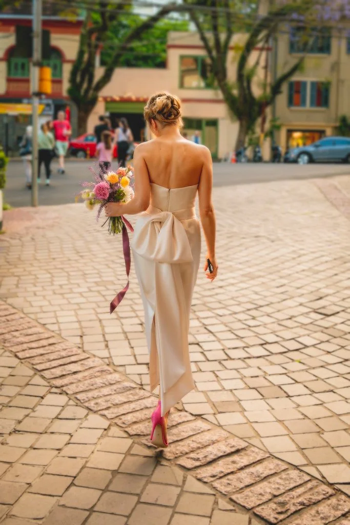 Mulher em vestido de festa calçando sapatos pink, carregando buquê de flores, caminhando na rua