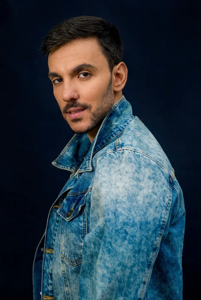 Homem com cabelo escuro e barba, usando jaqueta jeans azul, olhando para a câmera, fundo escuro