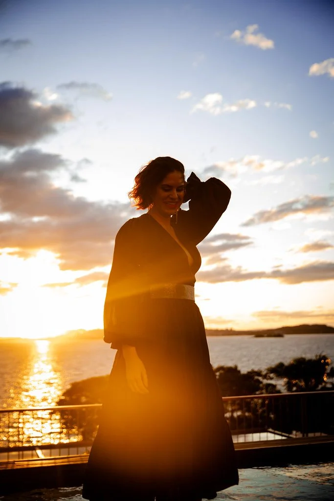 Mulher sorridente ao pôr do sol em frente a um rio, vestindo vestido longo escuro.