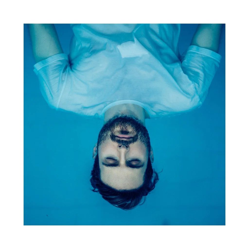 Homem com barba, de olhos fechados, deitado na água com camiseta branca.