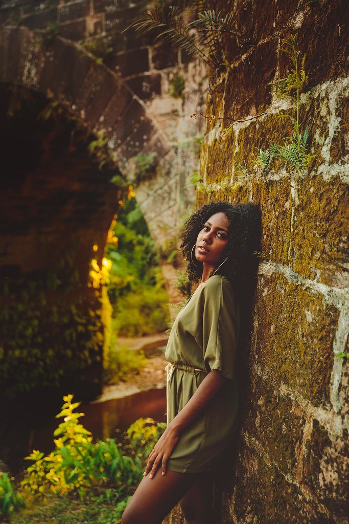Mulher jovem com cabelo cacheado, usando vestido verde, apoiada em parede de pedra com vegetação ao redor, ao pôr do sol.