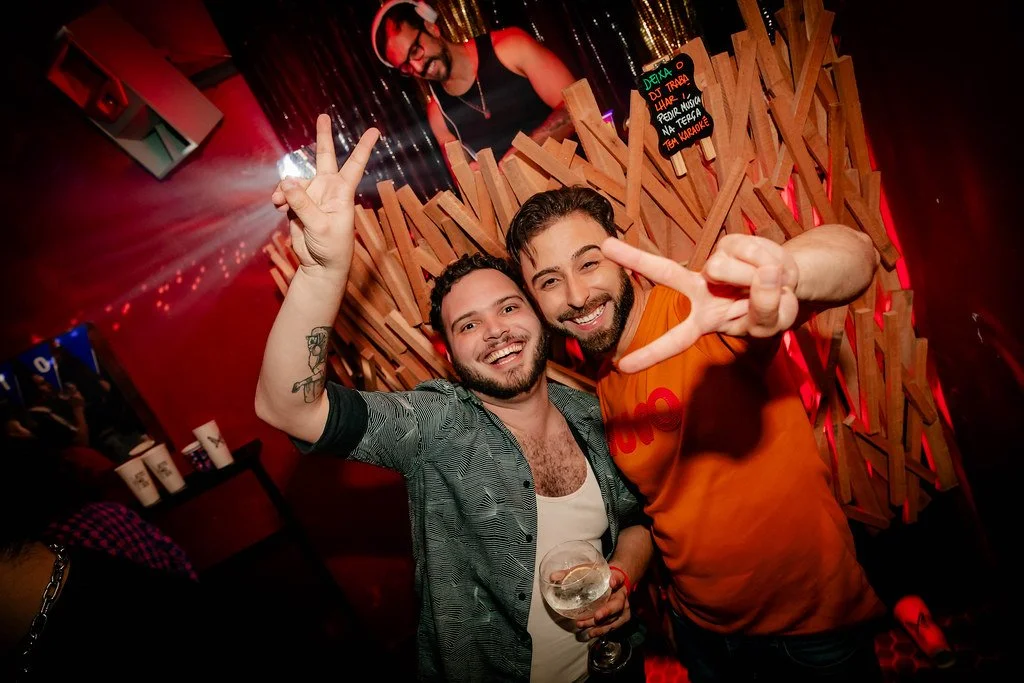 Dois homens sorridentes fazendo sinal de paz com as mãos, em uma festa, com iluminação colorida e fundo decorado com painéis de madeira.