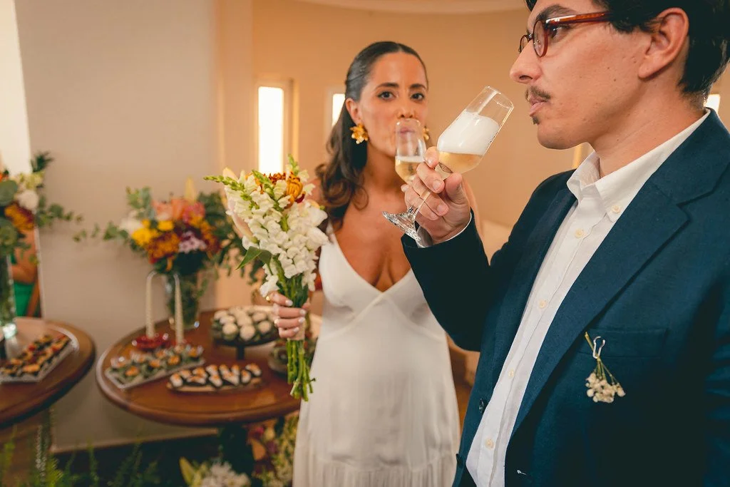 Casal brindando com champagne em uma comemoração de casamento, com mesa de doces ao fundo.