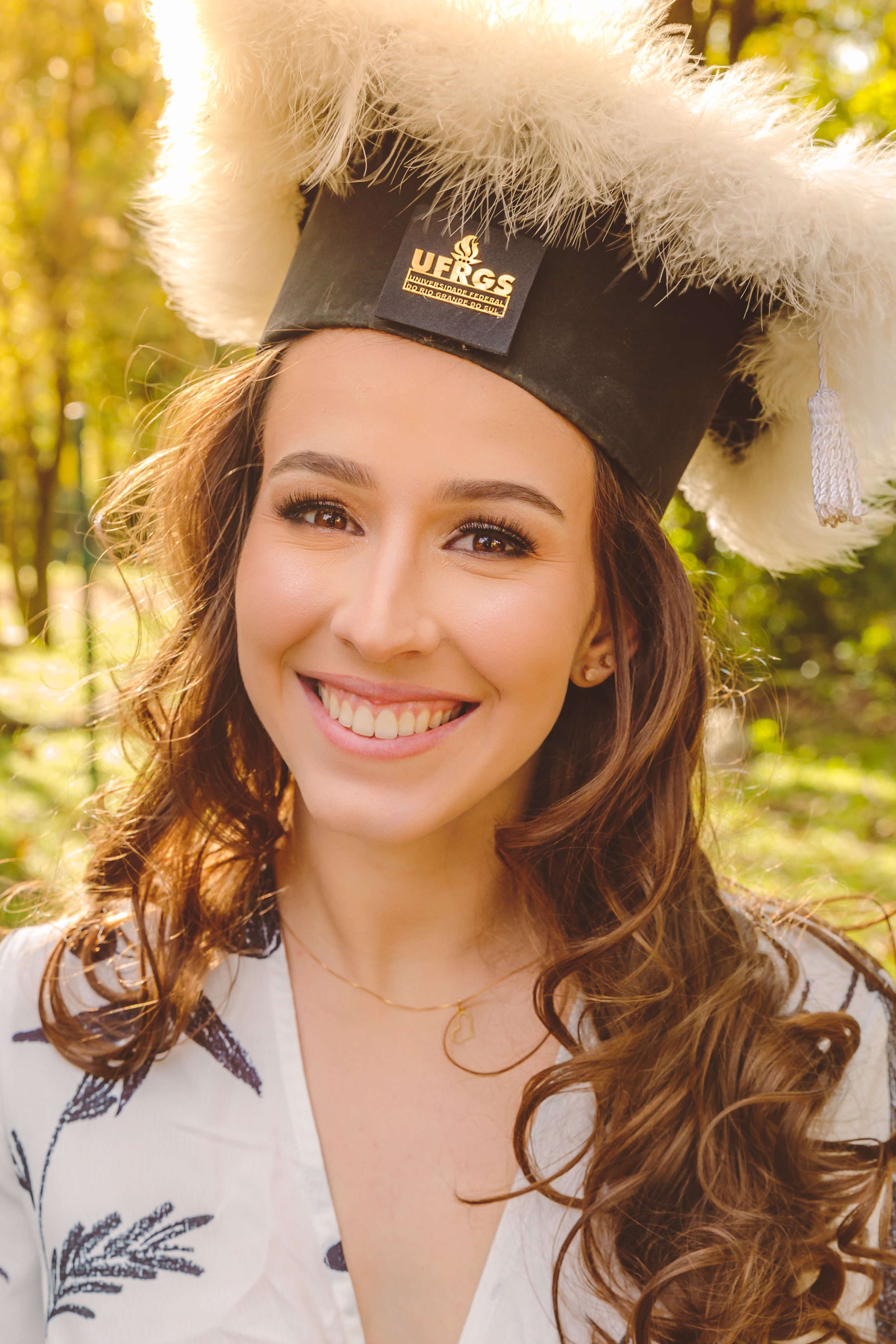 Mulher sorrindo com chapéu de formatura, cabelo castanho e ondulado, em fundo de natureza com árvores.