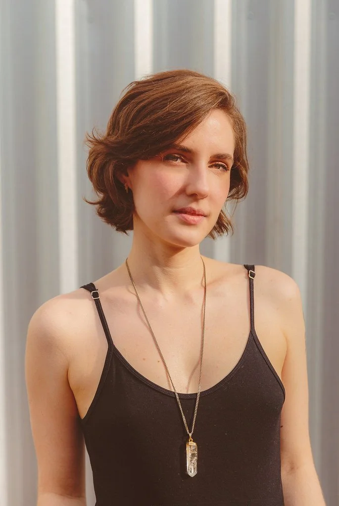 Mulher com cabelo castanho, usando camiseta preta sem mangas e um colar com pingente de cristal.