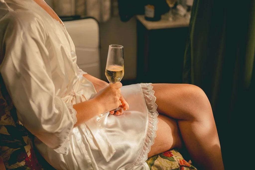 Pessoa usando roupão de seda branco, sentada, segurando uma taça de champanhe, com as pernas cruzadas.