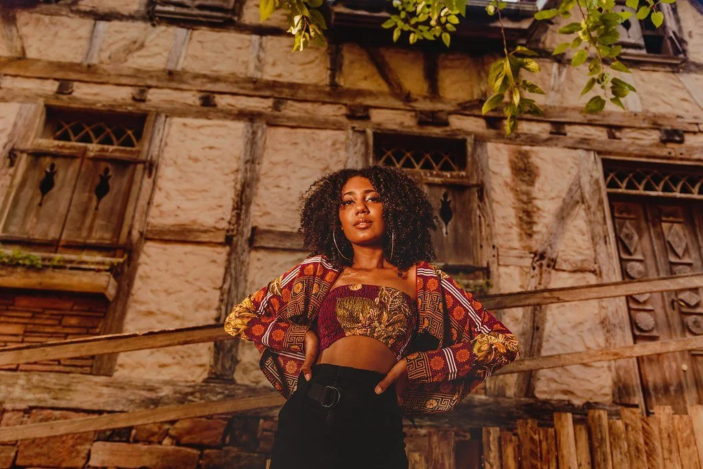Mulher jovem com cabelo cacheado, vestindo uma blusa de manga longa colorida e um top de crochê, posando em frente a uma casa de madeira antiga com janelas de madeira decorativas.