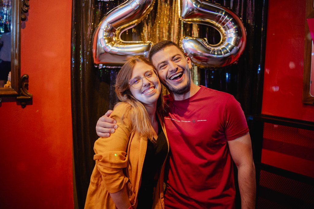 Homem e mulher sorrindo em comemoração, abraçados, com balões em formato de coração ao fundo.
