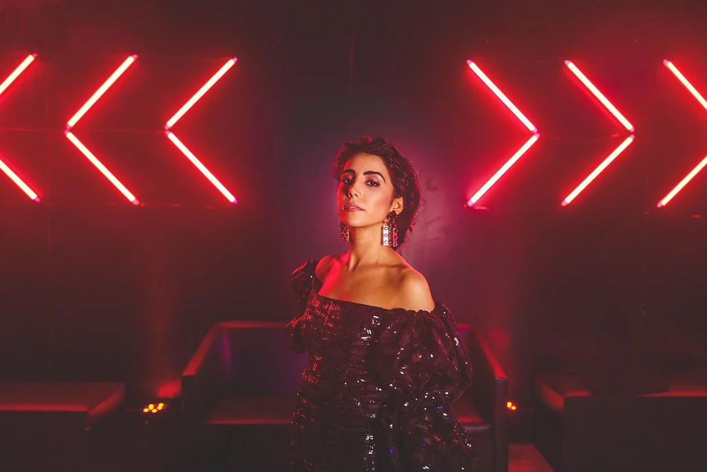 Mulher com vestido preto brilhante e brincos grandes, de cabelo escuro e penteado, posando em ambiente com iluminação de neon vermelho formando setas apontando para a direita, ao fundo.