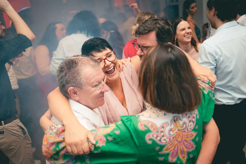 Grupo de pessoas se abraçando e sorrindo em uma festa, ambiente animado com várias pessoas ao fundo.