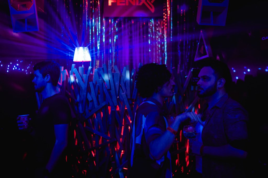 Três pessoas em uma festa com iluminação colorida, uma delas brincando com uma outra, enquanto uma terceira observa de longe ao lado de uma parede decorada com materiais de madeira e luzes.