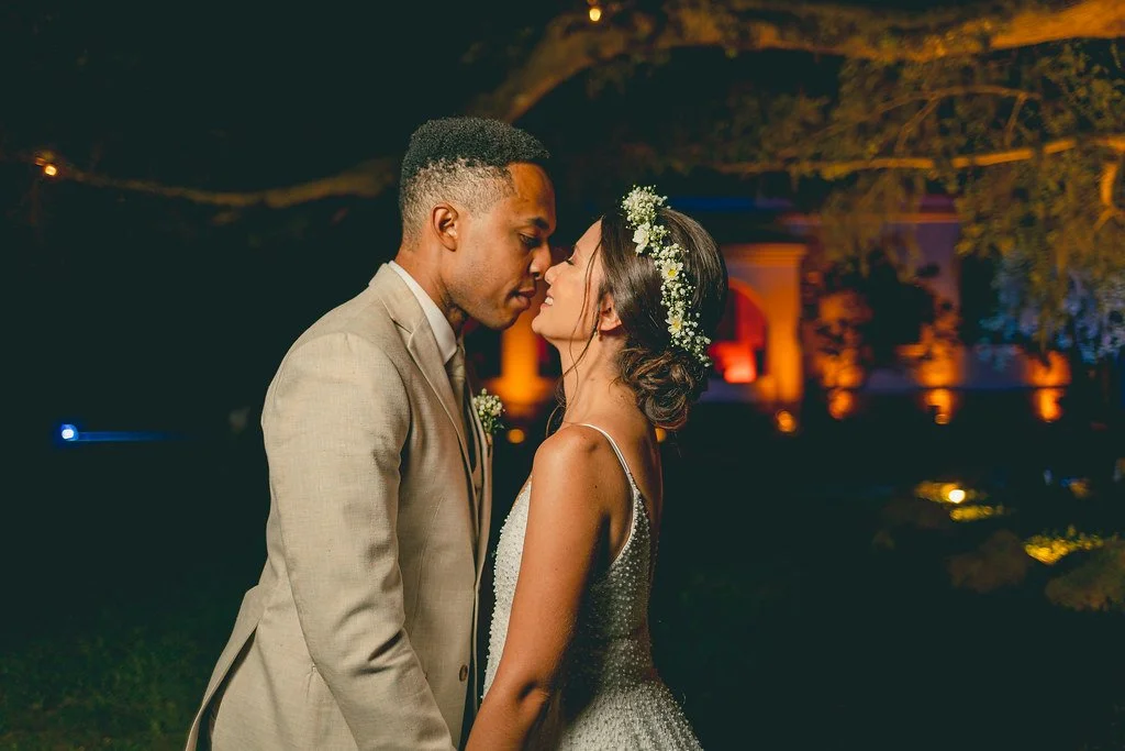 Casal de noivos no casamento, com o homem usando terno bege e a mulher usando vestido de noiva com tiara de flores, em um cenário ao ar livre à noite