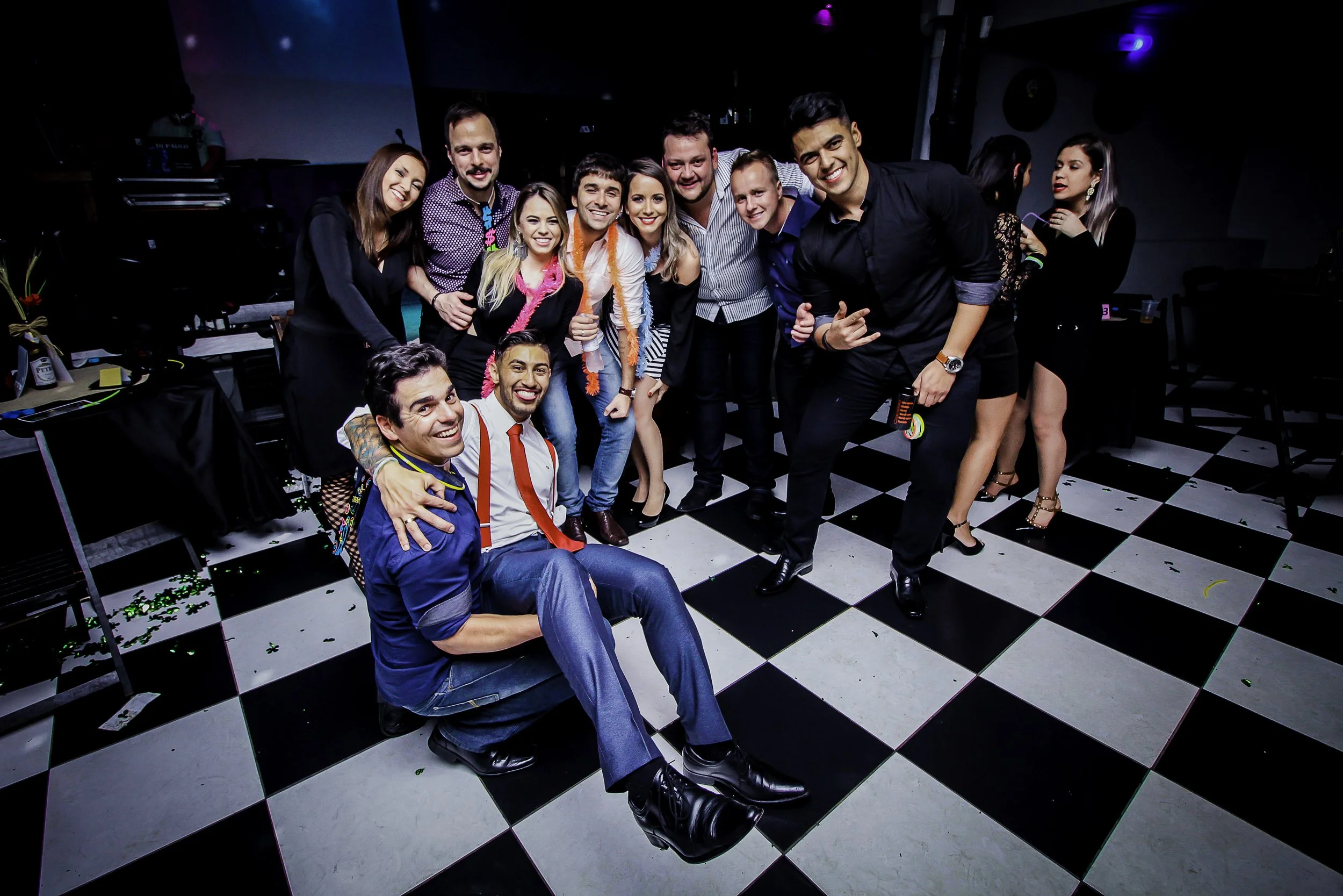 Grupo de pessoas comemorando em uma festa, sorrindo e posando em um ambiente com piso quadriculado preto e branco.