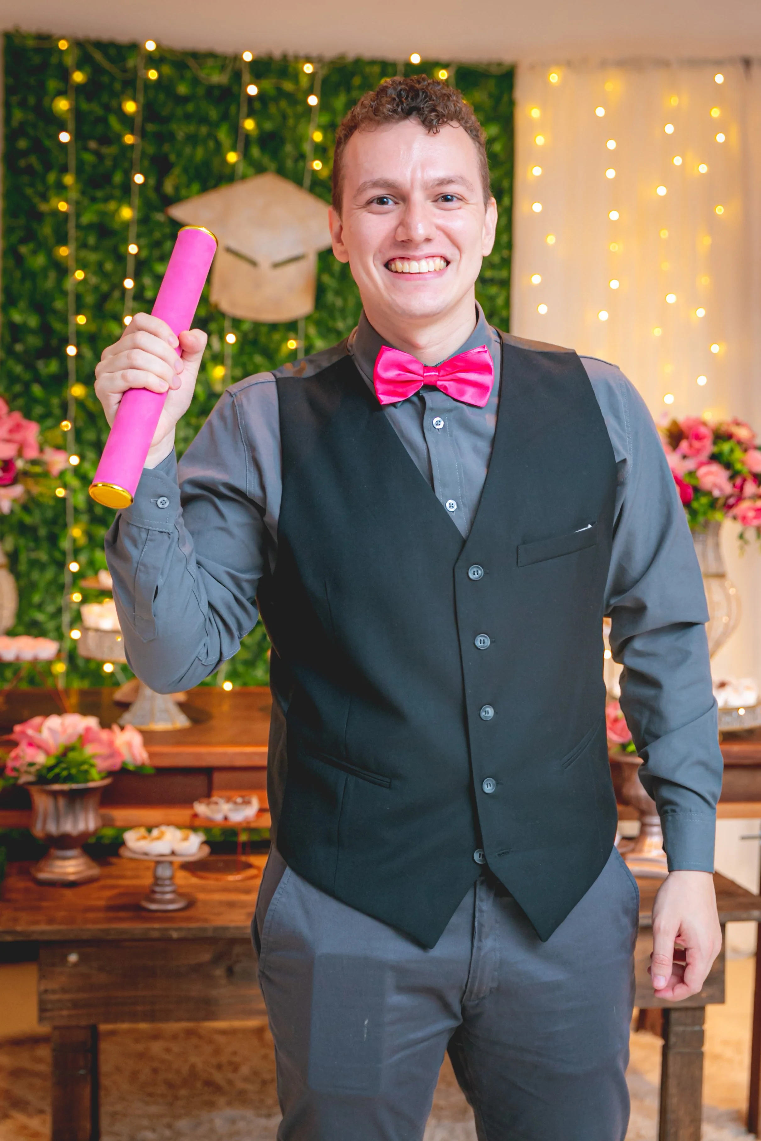 Homem sorridente vestido com colete escuro, camisa cinza, e gravata borboleta rosa, segurando um tubo rosa, em festa com decoração de luzes e flores ao fundo.