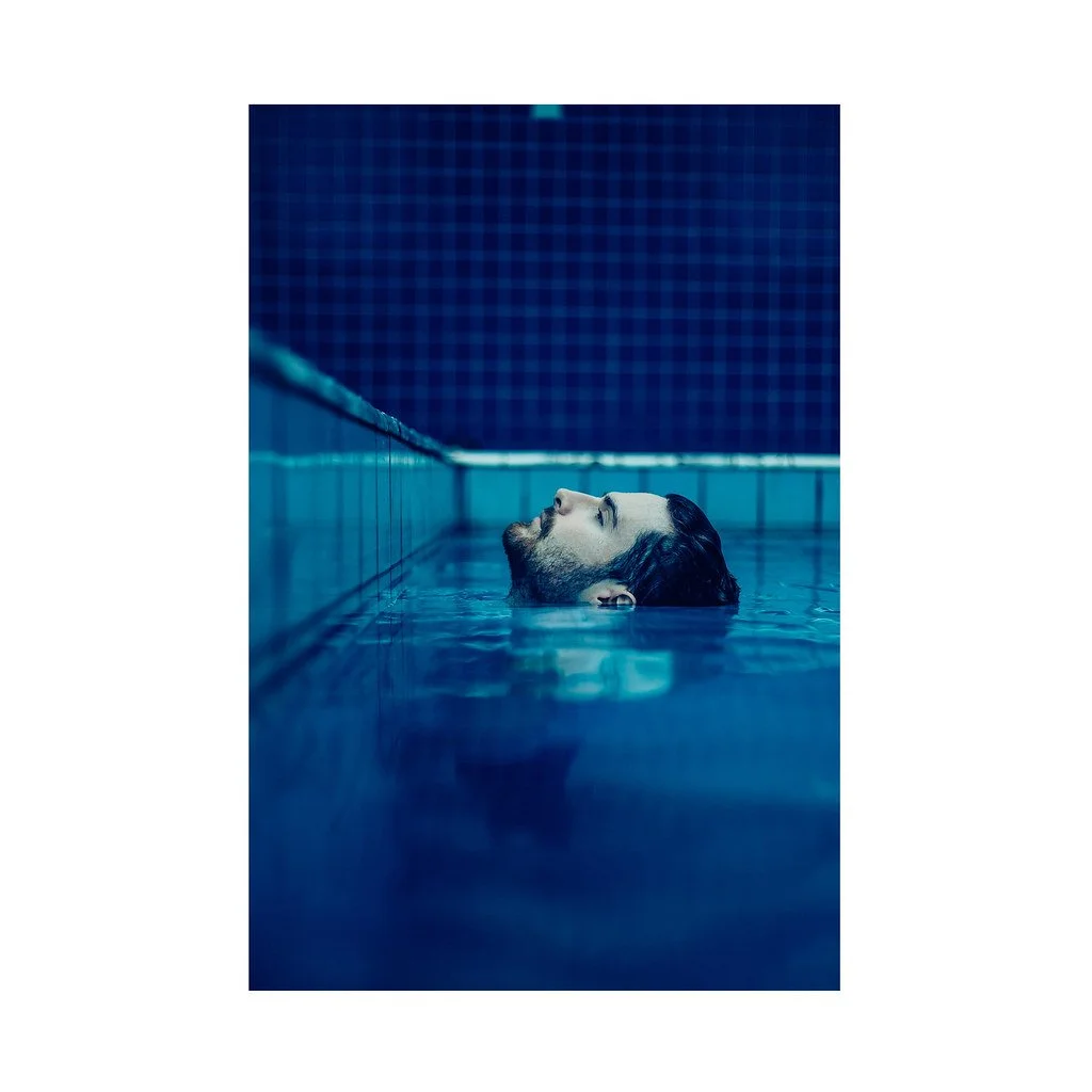 Homem com barba e cabelo escuro relaxando na piscina, com a cabeça apoiada na borda, nadando de costas, numa piscina com azulejos azuis.