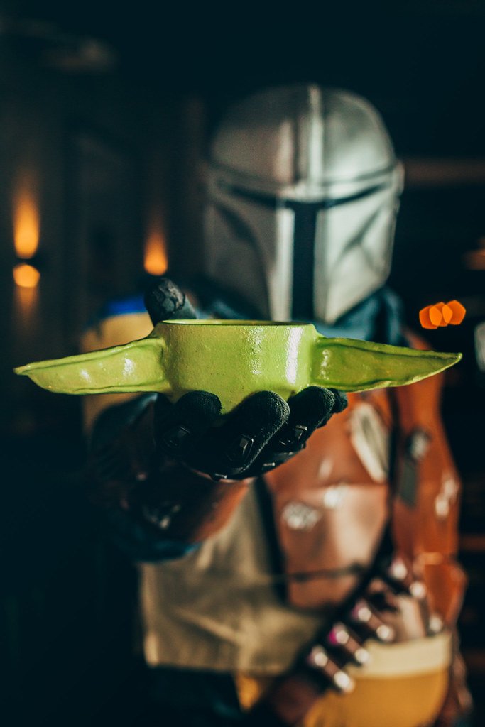Personagem de Star Wars Barberança usando capacete branco e roupa marrom, segurando um prato com um pedaço de fruta em forma de orelhas de Baby Yoda.