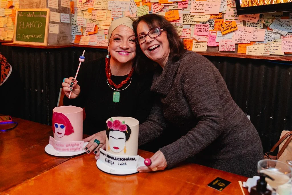 Duas mulheres sorridentes segurando dois bolos decorados com figuras femininas e textos. Estão em uma mesa de restaurante com parede de post-its ao fundo.