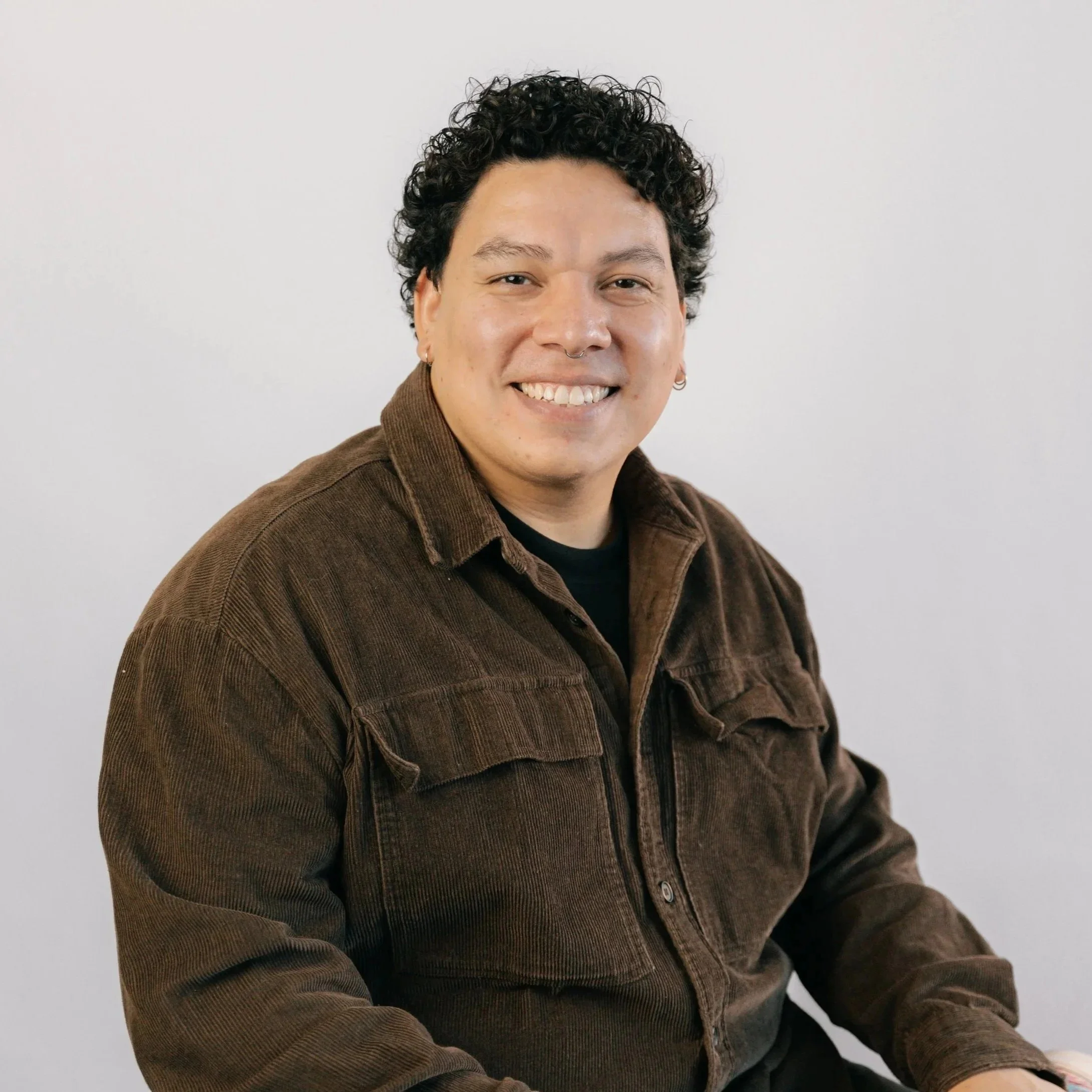 Retrato de uma pessoa sorrindo, com cabelo cacheado escuro, usando uma jaqueta marrom, contra fundo branco.