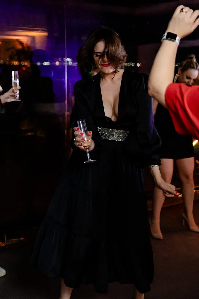 Mulher usando vestido preto longo, com cabelo ondulado, segurando uma taça de champanhe em um ambiente de festa à noite.