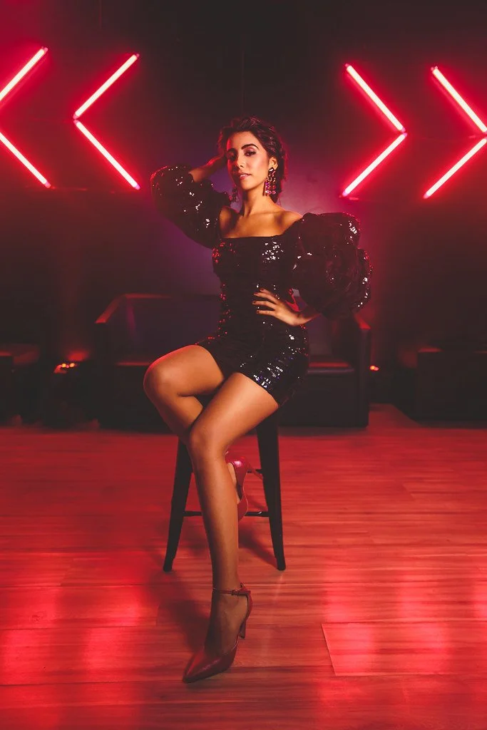 Mulher vestida com vestido preto brilhante, sentado em uma cadeira, com cabelo escuro, brincos grandes, posando com uma mão na cabeça e a outra na cintura, em ambiente com iluminação neon vermelha.