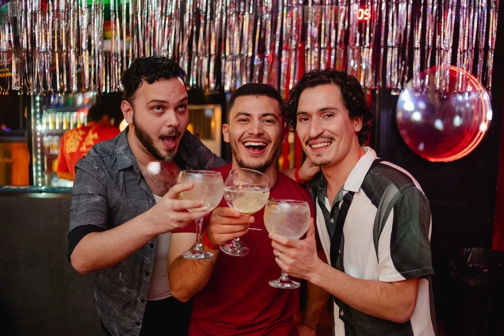 Três homens sorrindo e brindando com coquetéis em uma festa, decorada com fita metálica e uma bola de espelho.