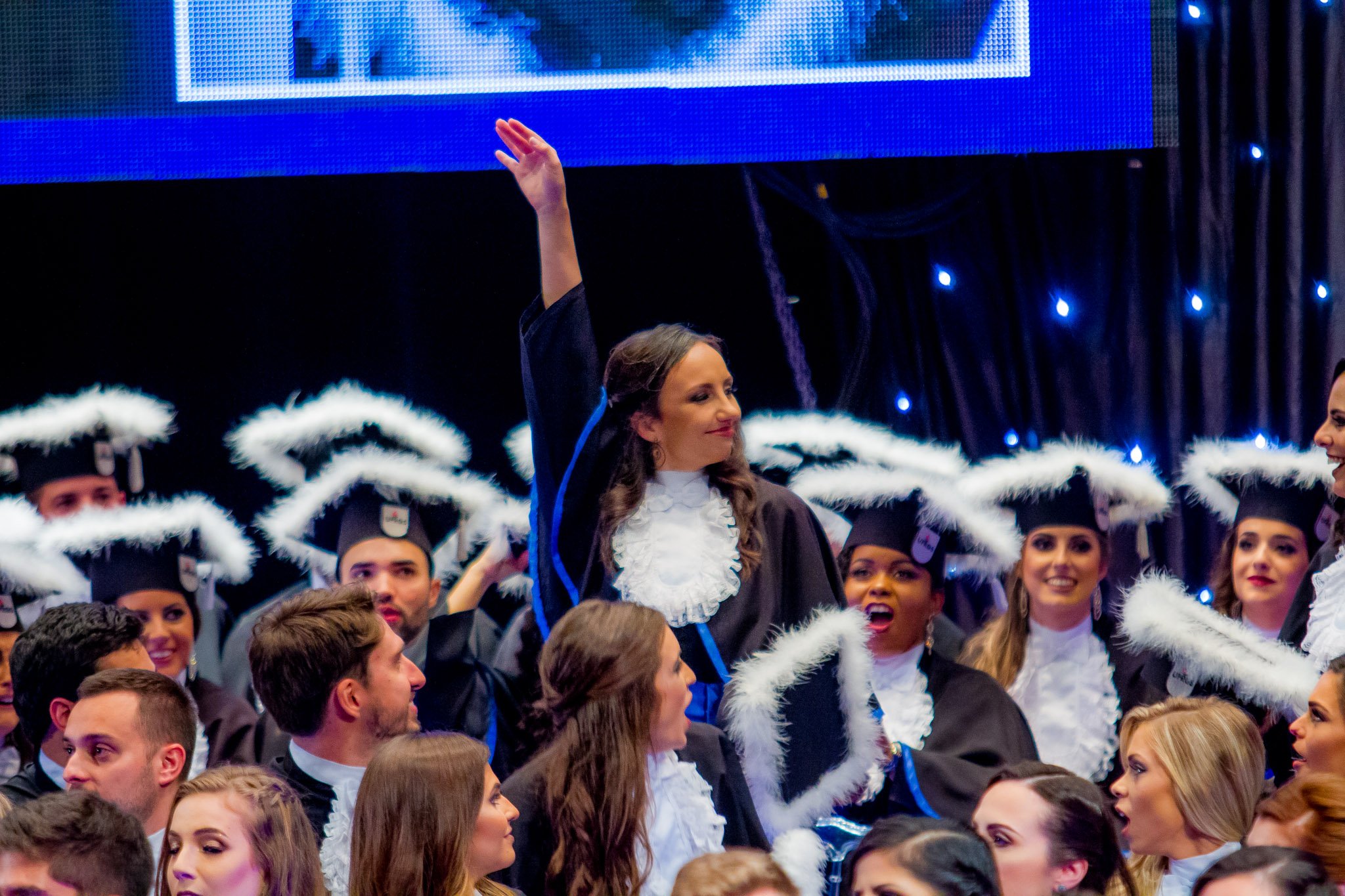 Muitos estudantes de graduação em trajes acadêmicos, incluindo becas pretas com detalhes brancos e chapéus com friséis brancos, celebram em uma cerimônia de formatura, com uma mulher no centro levantando a mão.