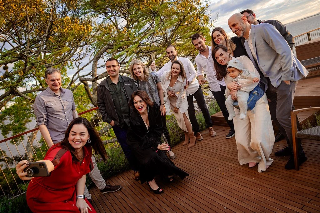 Grupo de pessoas sorrindo e posando para uma foto ao ar livre, em um deck de madeira com árvores ao fundo, durante o dia.