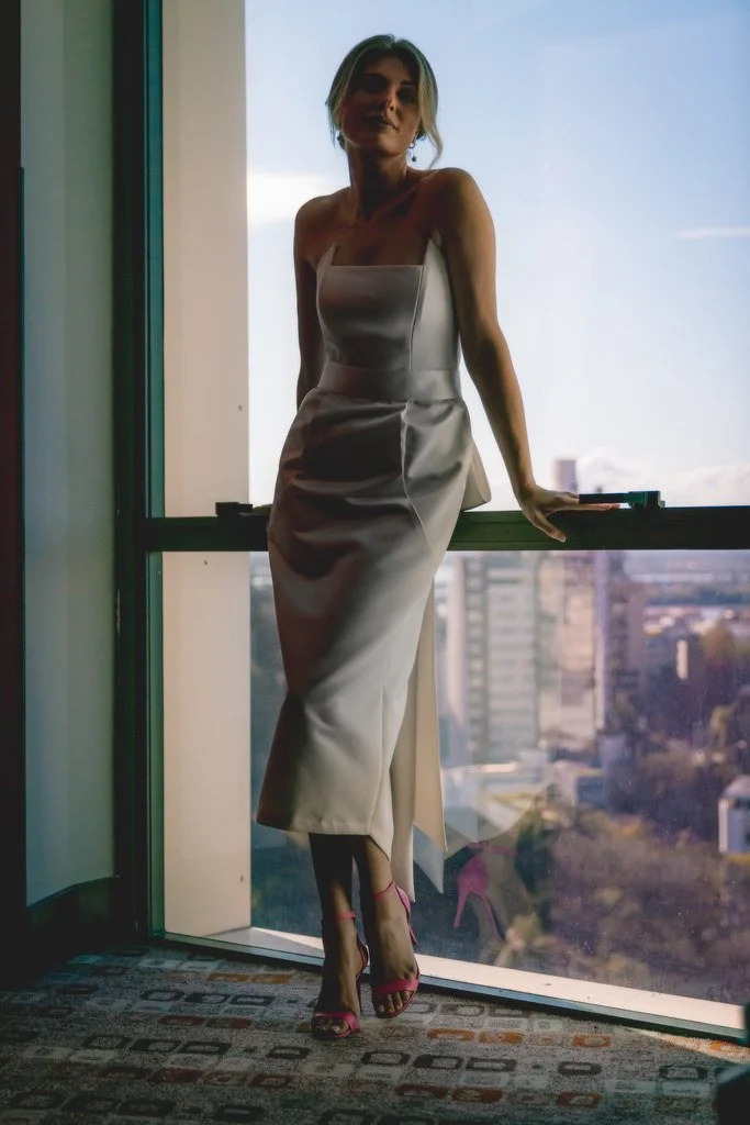 Mulher de vestido longo sentada na janela de um prédio com vista para uma cidade ao fundo.