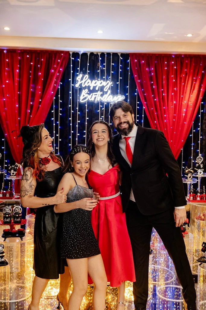 Grupo de pessoas comemorando em festa de aniversário com decoração de luzes, cortinas vermelhas e painel com mensagem "Happy Birthday" ao fundo.