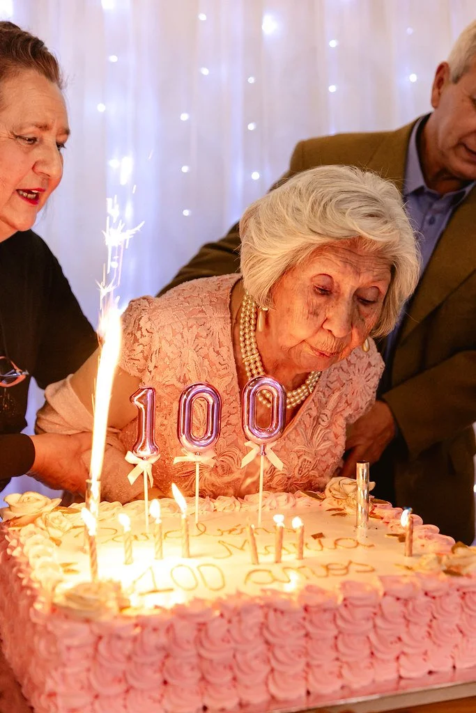 Mulher idosa celebrando 100 anos de idade em festa com bolo decorado e velas em forma de números 100.