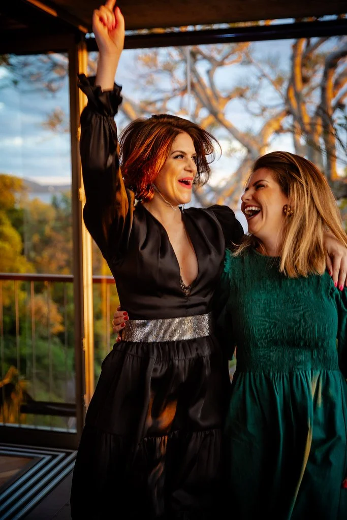 Duas mulheres sorrindo e brincando em um ambiente ao ar livre, uma de cabelo castanho-avermelhado usando vestido preto com faixa prateada, a outra de cabelo castanho claro usando vestido verde, com árvores ao fundo.