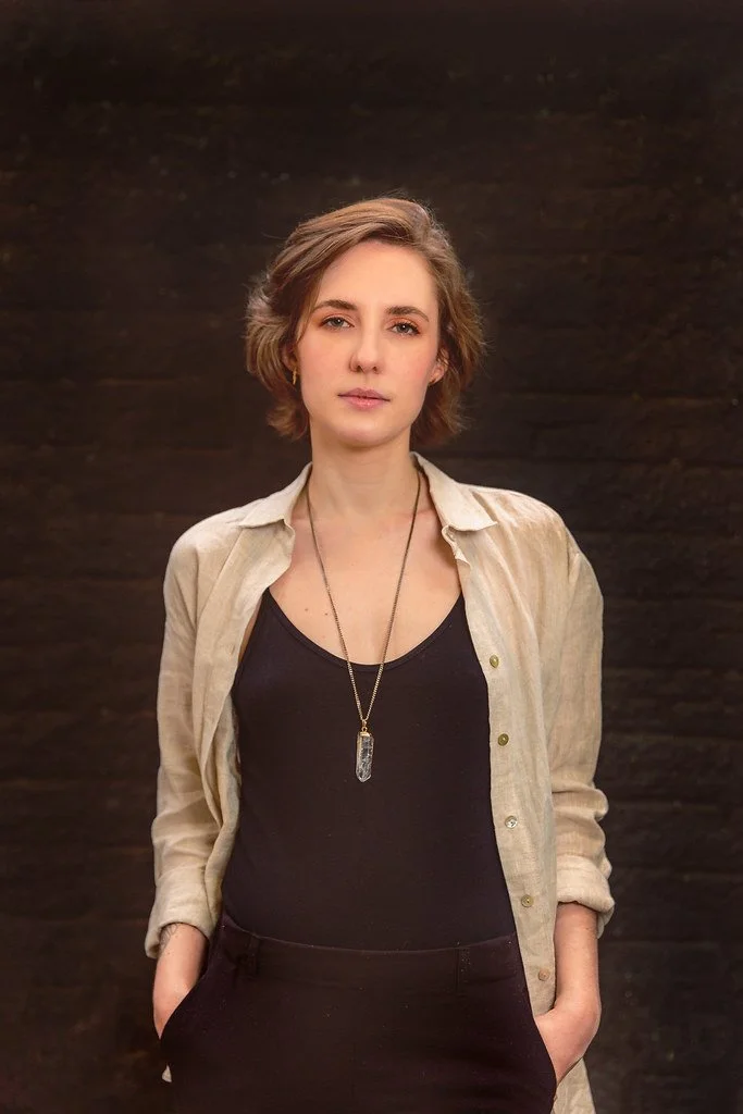 Retrato de uma jovem mulher com cabelo castanho curto, usando uma blusa preta, uma camisa bege aberta e um colar com pingente, posando em frente a uma parede escura.