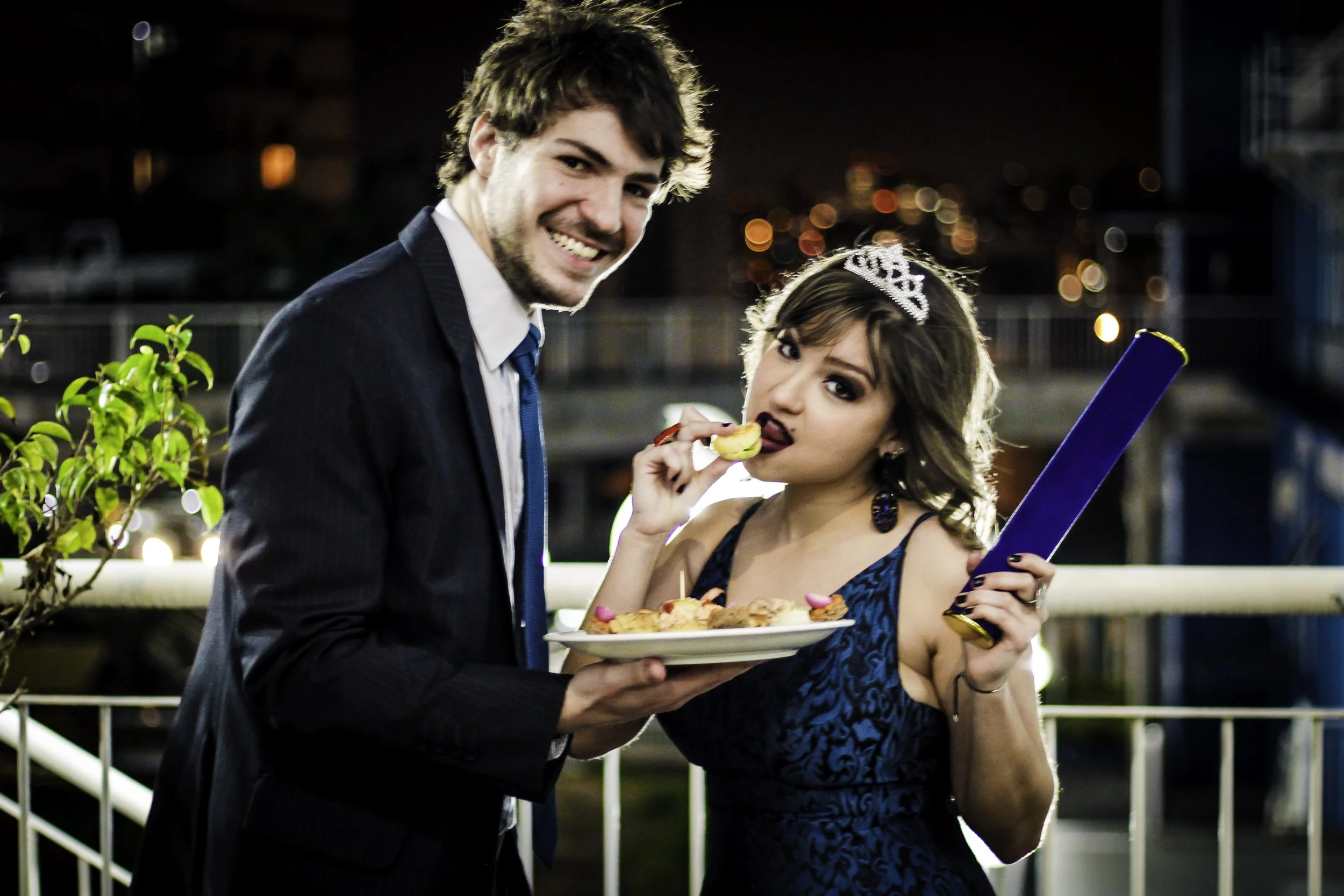 Casal celebrando uma festa de casamento à noite, com o homem segurando um prato de sobremesas e a mulher usando uma tiara, com uma placa azul na mão, em um ambiente ao ar livre na cidade.