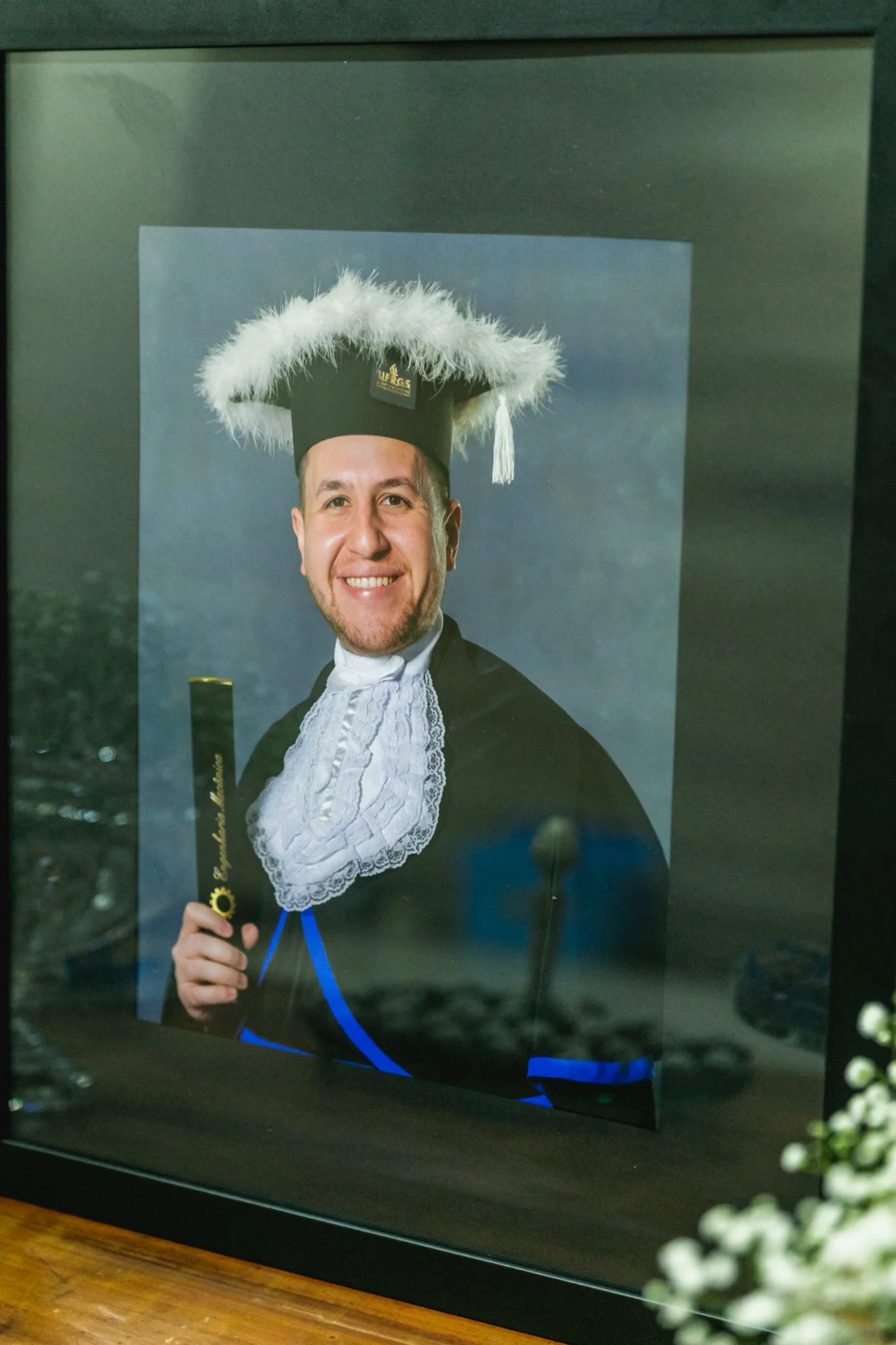 Fotografia de um homem sorridente vestido com roupa de formatura, usando beca preta, toca com vistoso adorno branco, e uma medida na mão, possivelmente de formatura.