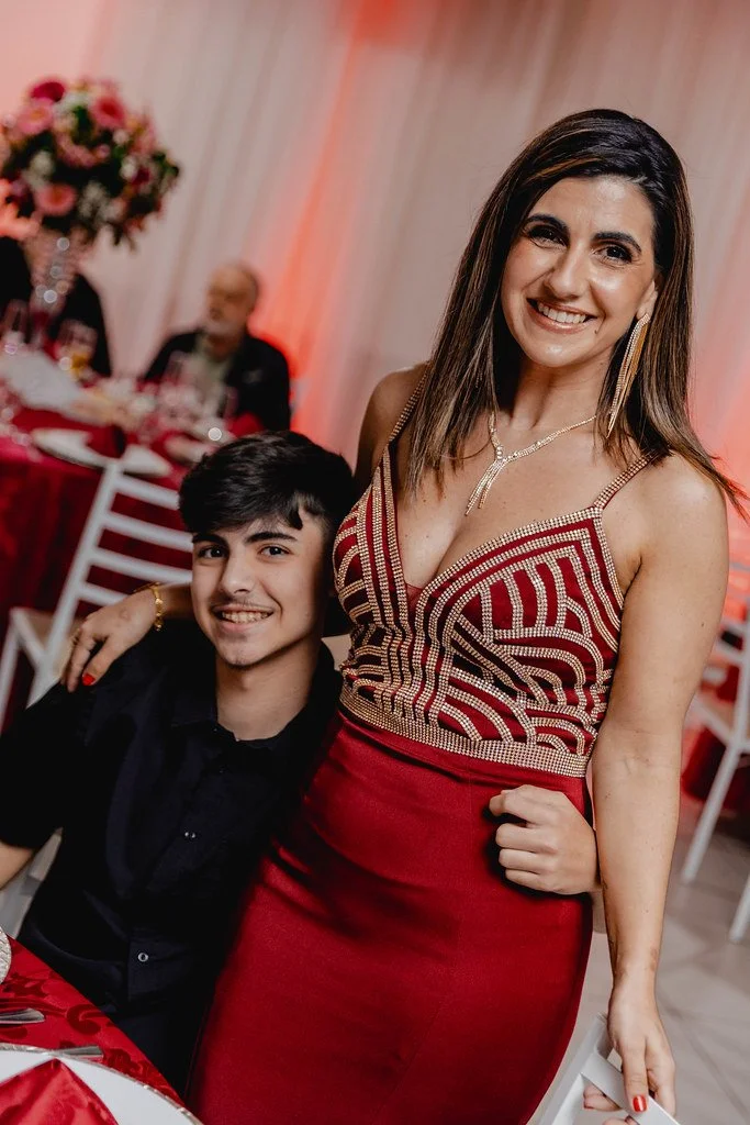 Mulher sorridente posando ao lado de um jovem rapaz sentado em uma festa, com decoração em tons de vermelho e flores ao fundo.