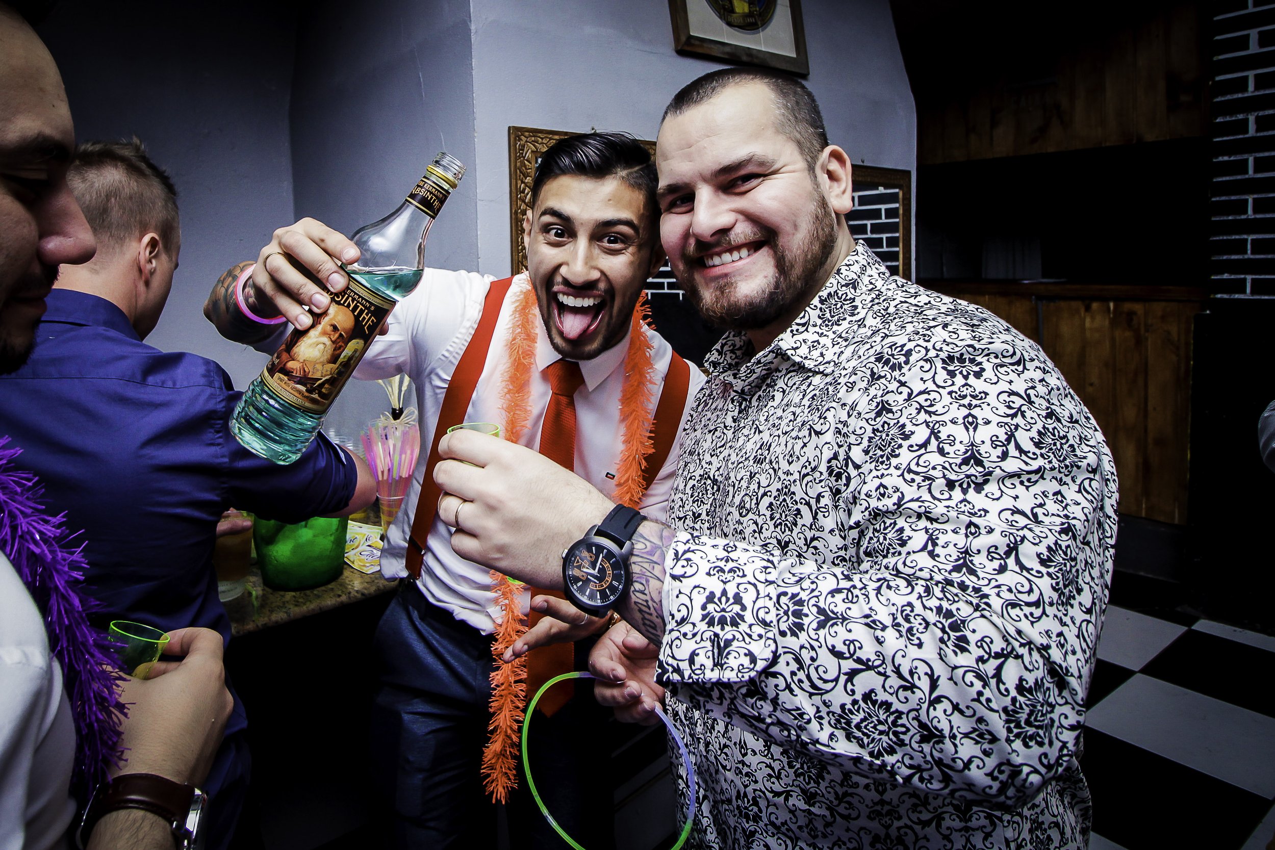 Homem com camisa de manga longa de padrão floral, usando relógio, e outro homem com gravata vermelha, usando uma faixa de pena laranja, ambos sorrindo e brindando em uma festa com outras pessoas ao fundo.