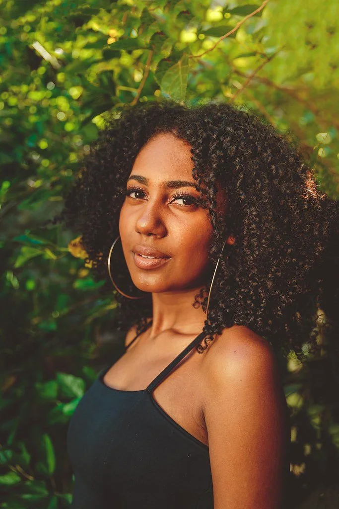 Retrato de uma mulher negra com cabelos cacheados escuros, usando brincos grandes de argola e blusa preta, em ambiente ao ar livre com vegetação verde ao fundo.