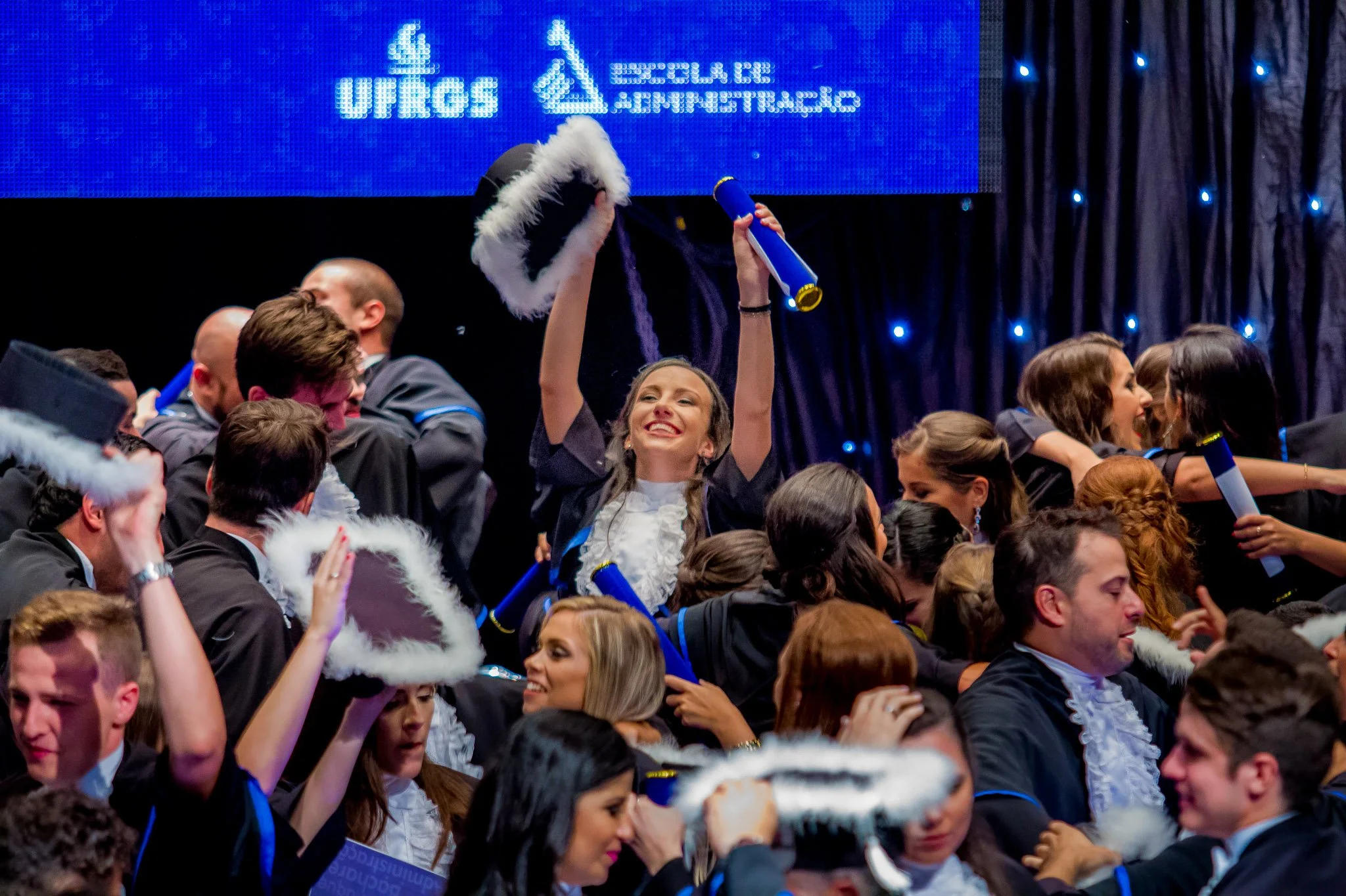 Grupo de pessoas comemorando em formatura, usando becas e chapéus, com uma mulher sorridente no centro levantando seu capelo e diploma. Fundo com tela digital e iluminação de palco.