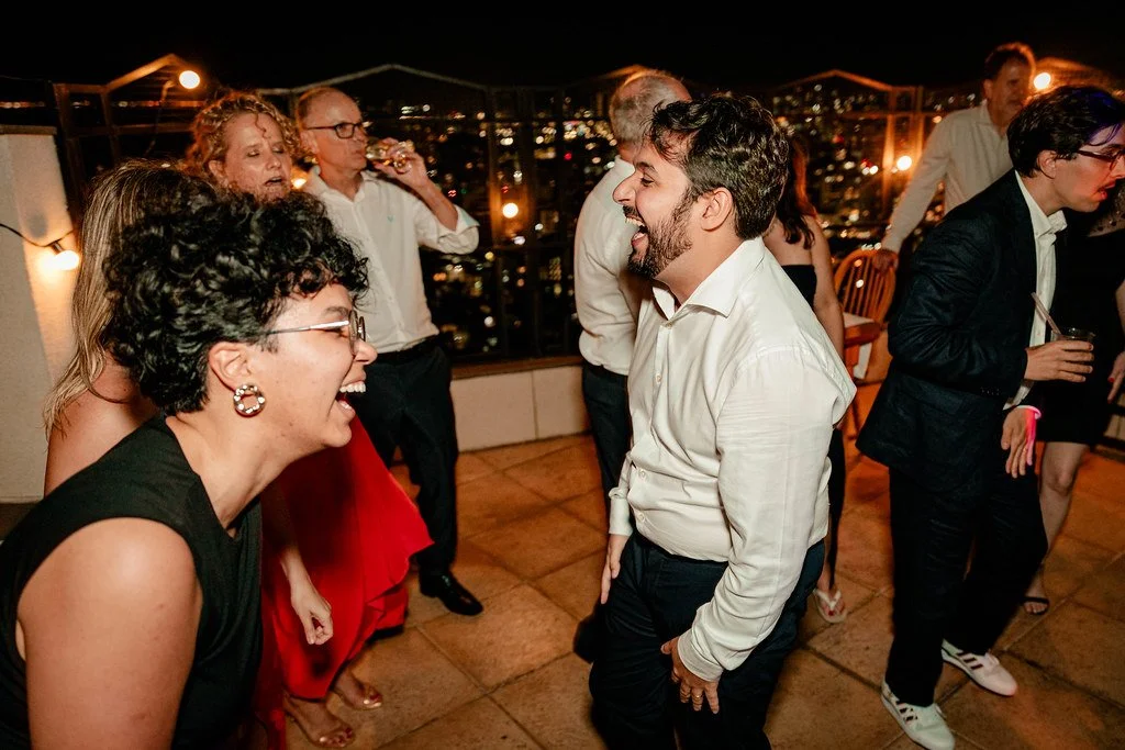 Grupo de pessoas sorrindo e conversando em uma festa noturna no terraço com vista para a cidade ao fundo.