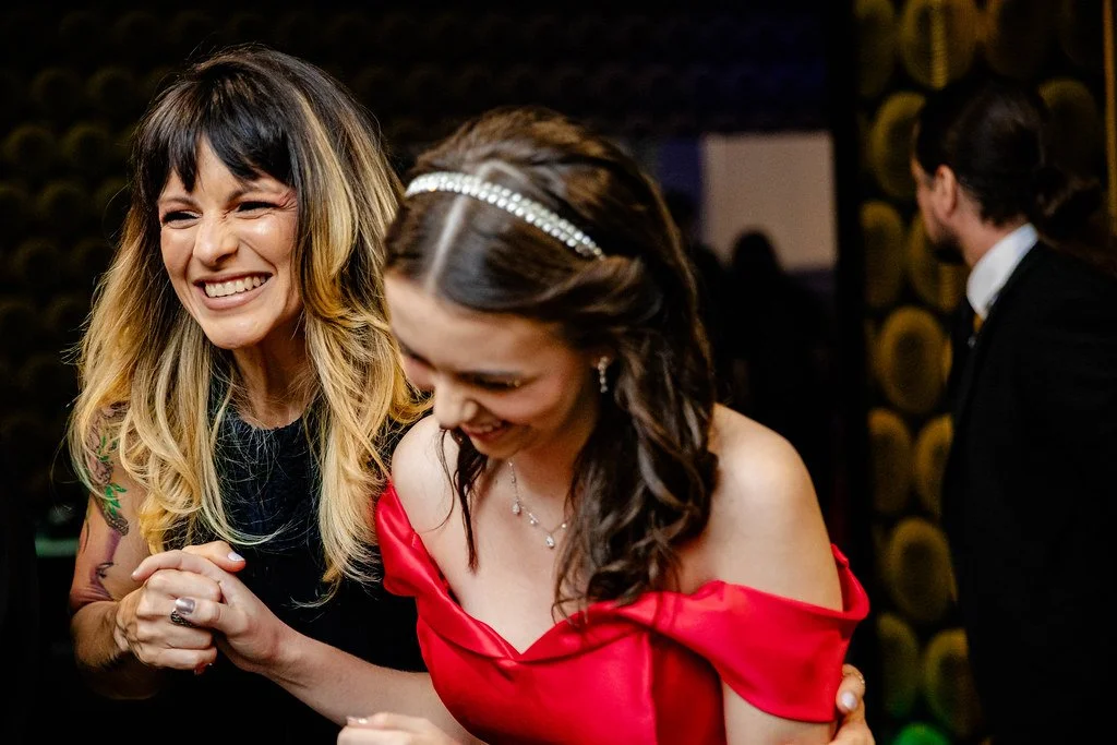 Duas mulheres sorrindo e dançando em uma festa, uma usando um vestido preto e a outra um vestido vermelho, em um ambiente festivo.