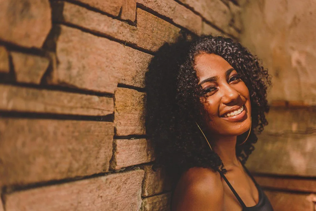 Mulher sorridente com cabelo cacheado ao lado de uma parede de tijolos.